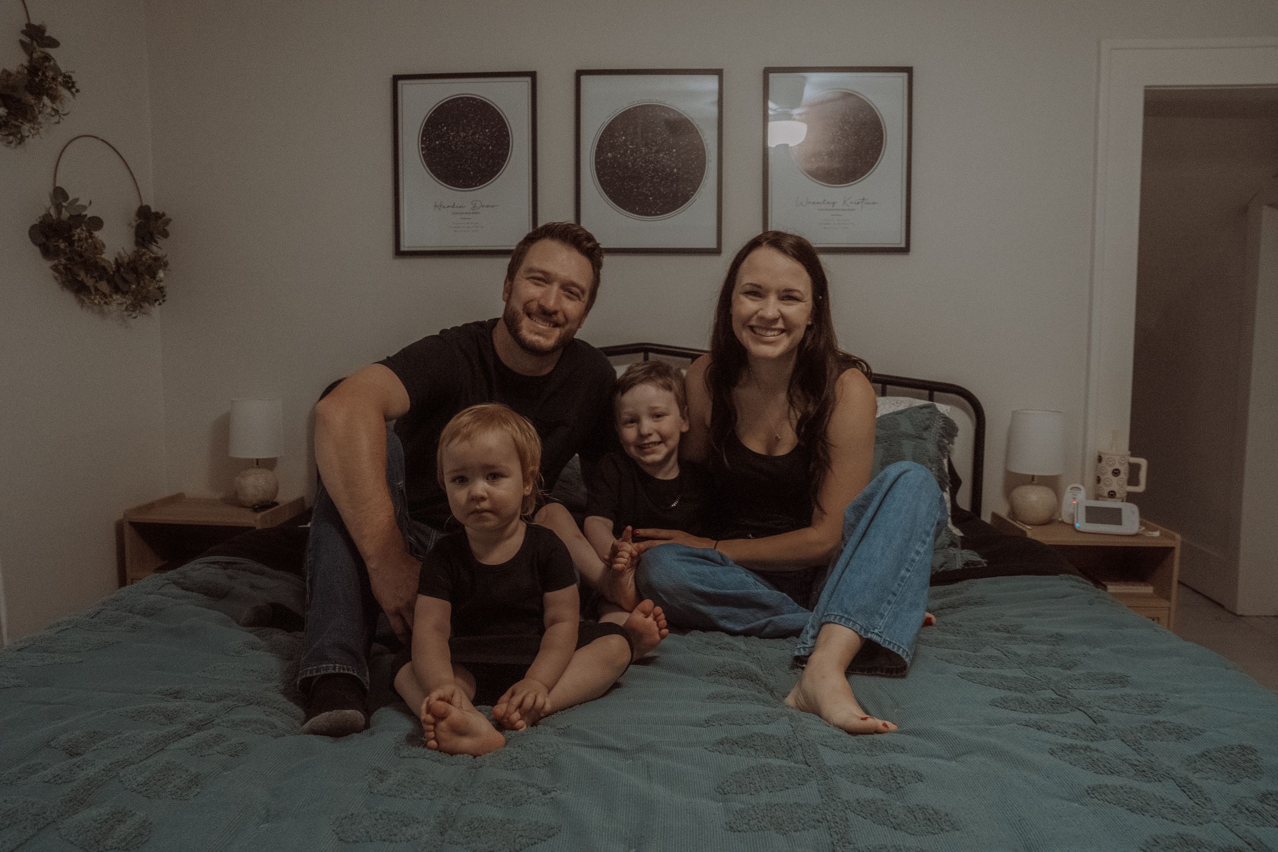 A family smiles together on a teal bedspread with lunar phase artwork hanging above them.
