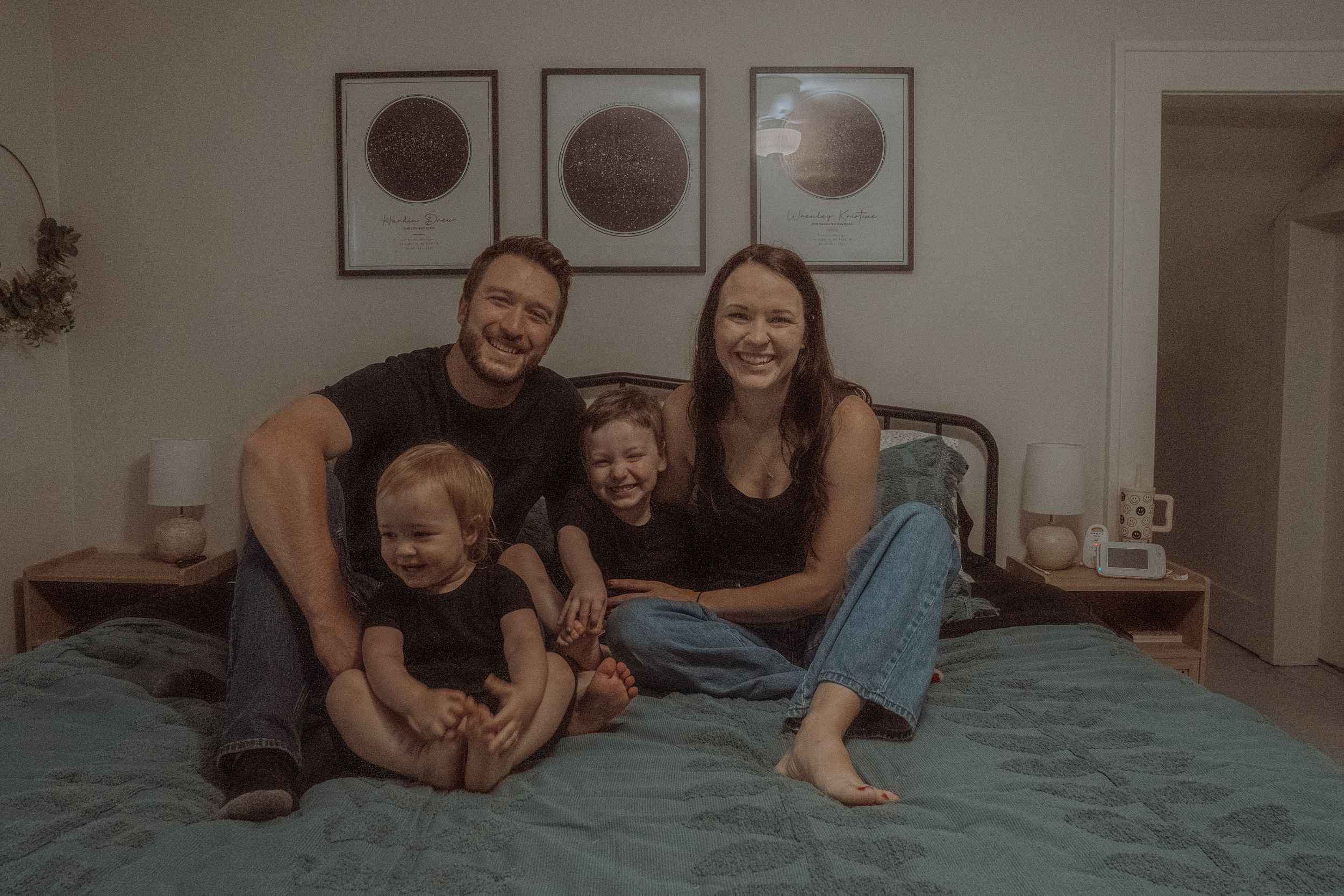 A family enjoys a playful moment together on a teal bed with moon phase artwork displayed on the wall.