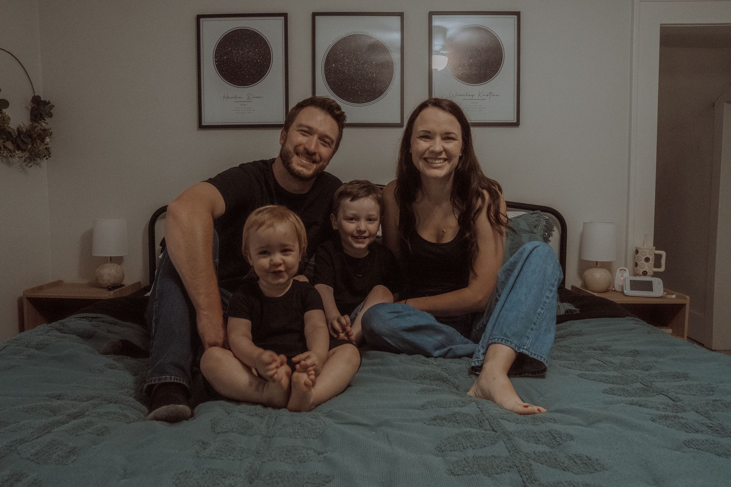 Candid family moment captured on bed with moon phase artwork displayed on wall above.