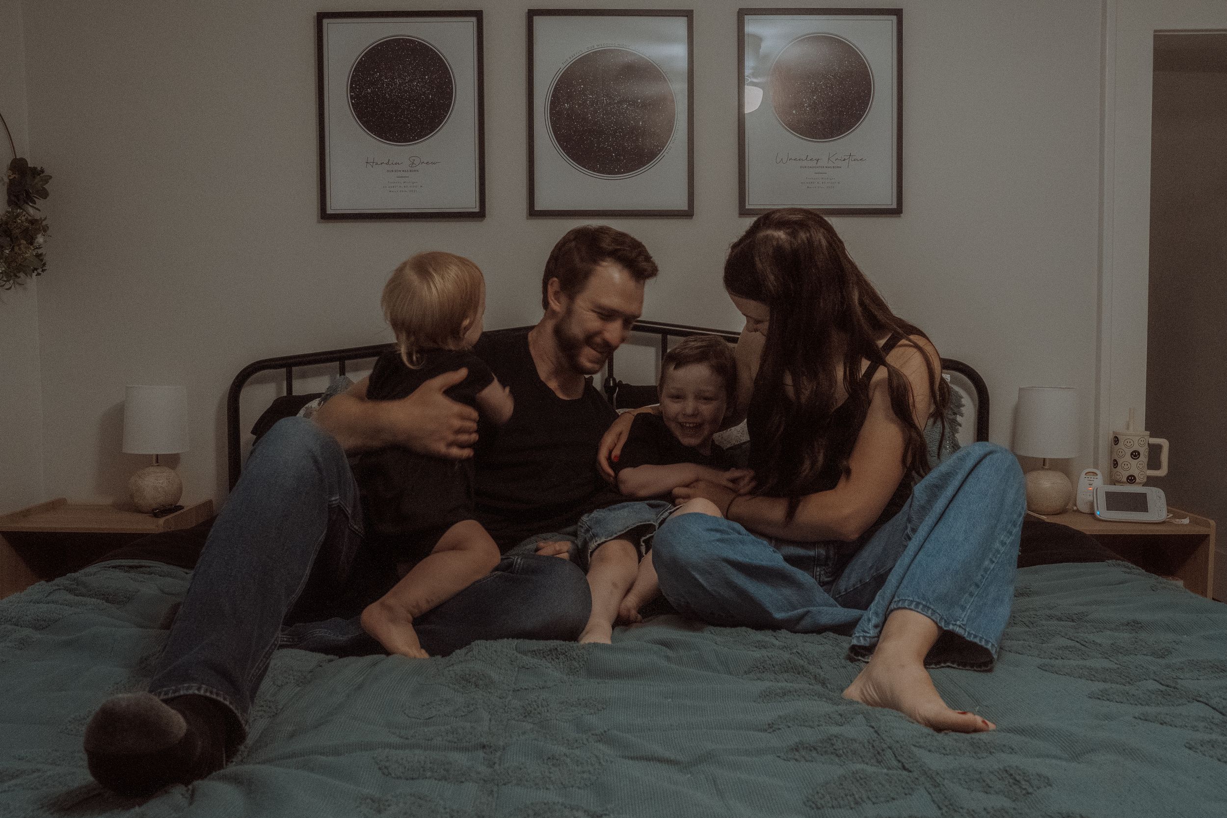 A family relaxes together on a teal bed in a cozy bedroom with moon phase artwork on the wall.