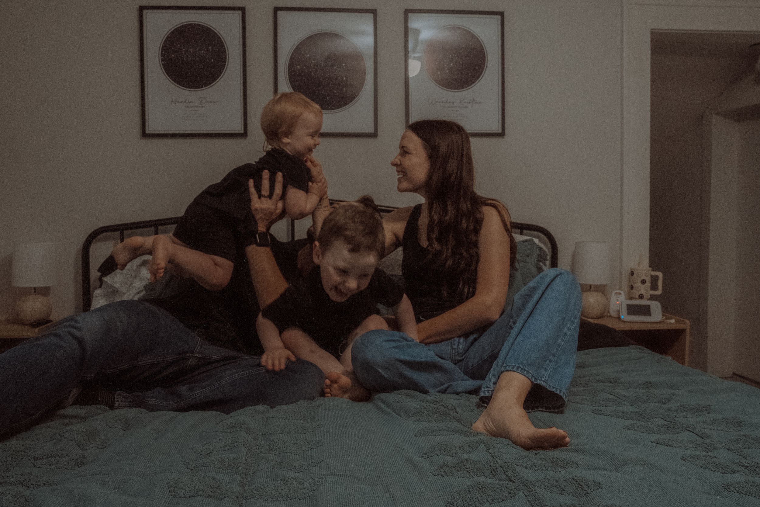 A couple playfully interacts with their child on a teal bed with moon phase artwork displayed on the wall.