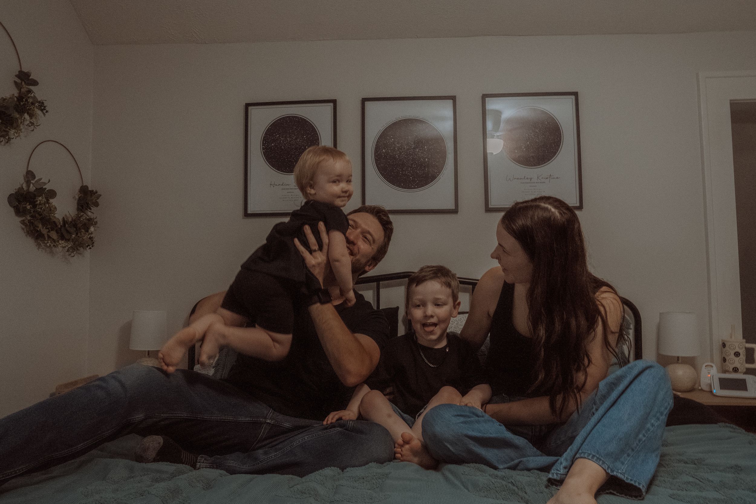 A candid moment of a family laughing and playing together on a bed with moon phase artwork in the background.
