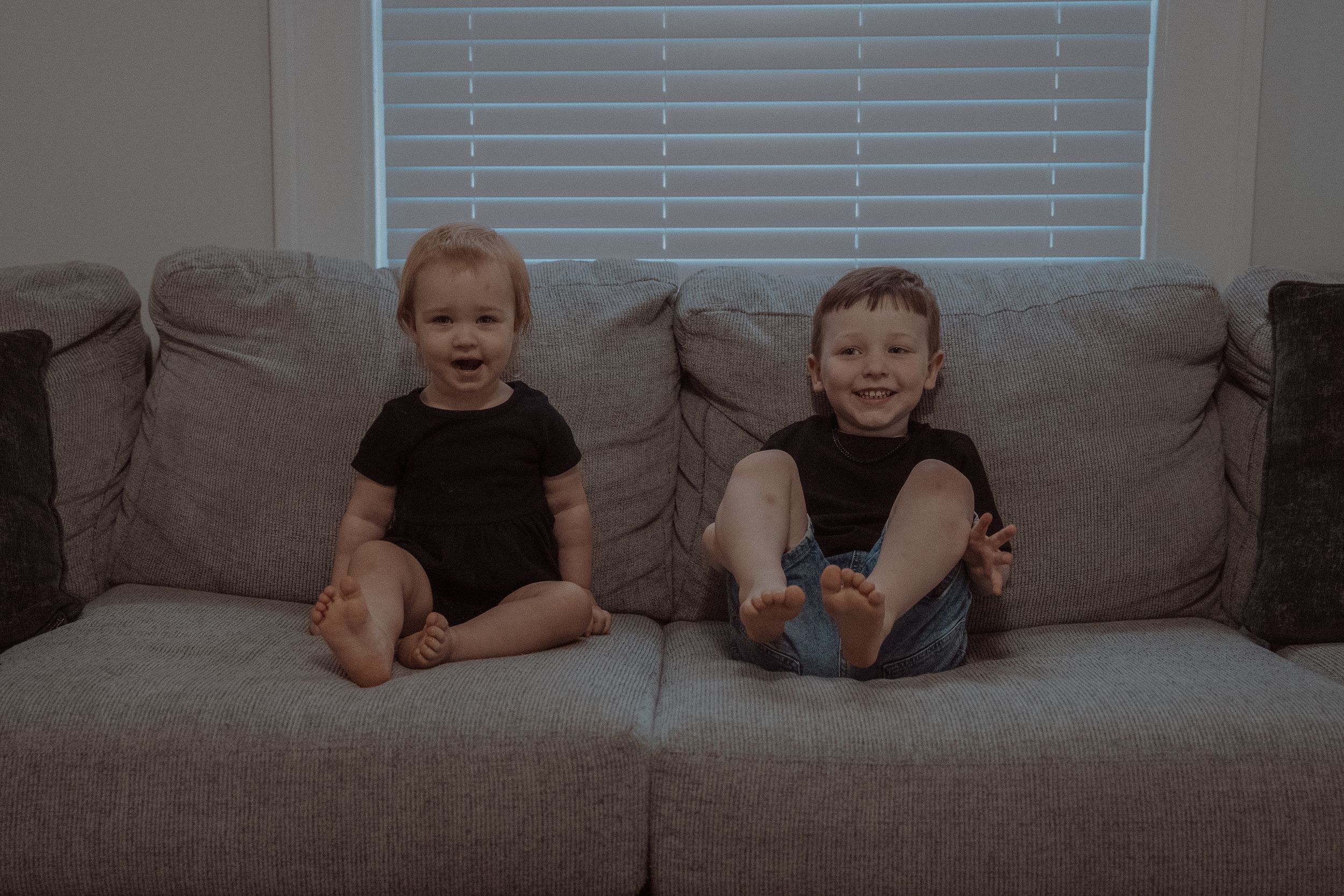 Two young children sit together on a gray couch in front of white venetian blinds.