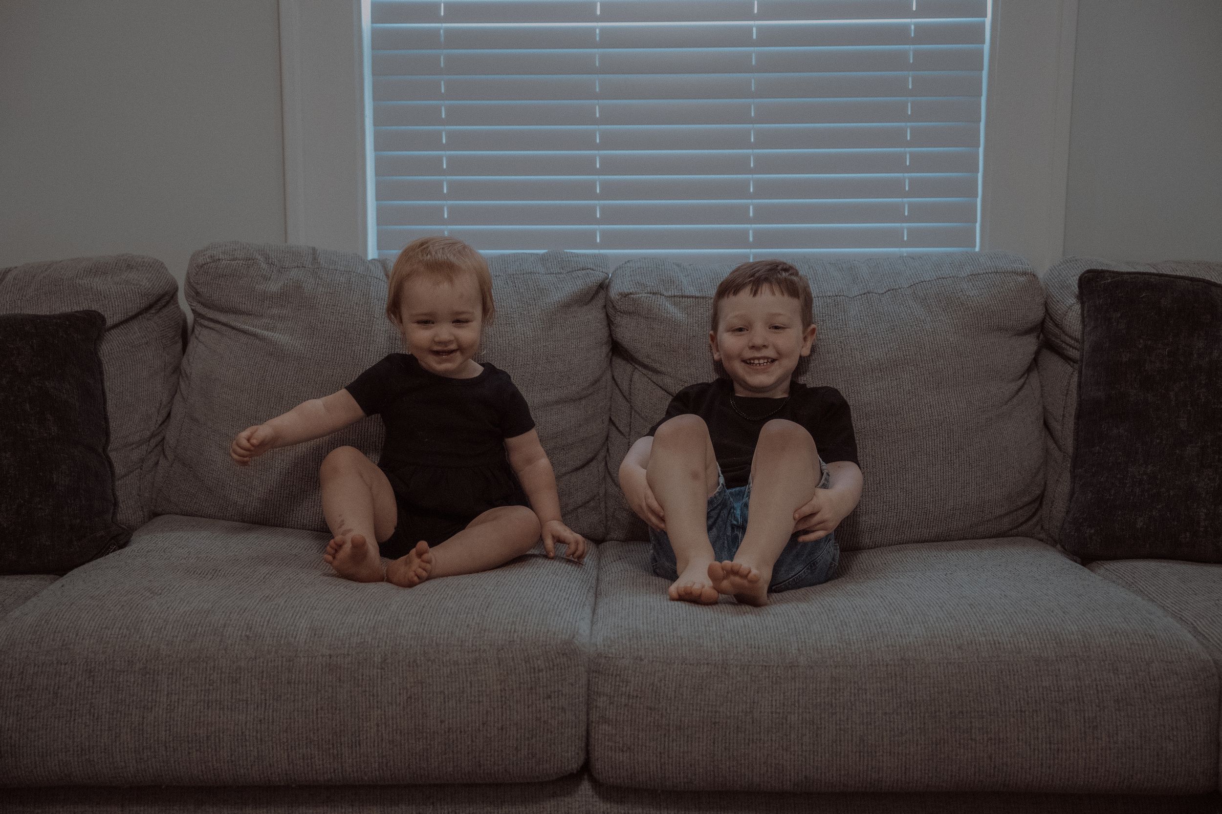 Two children lounge and laugh together on a gray sectional couch in a casual home setting.