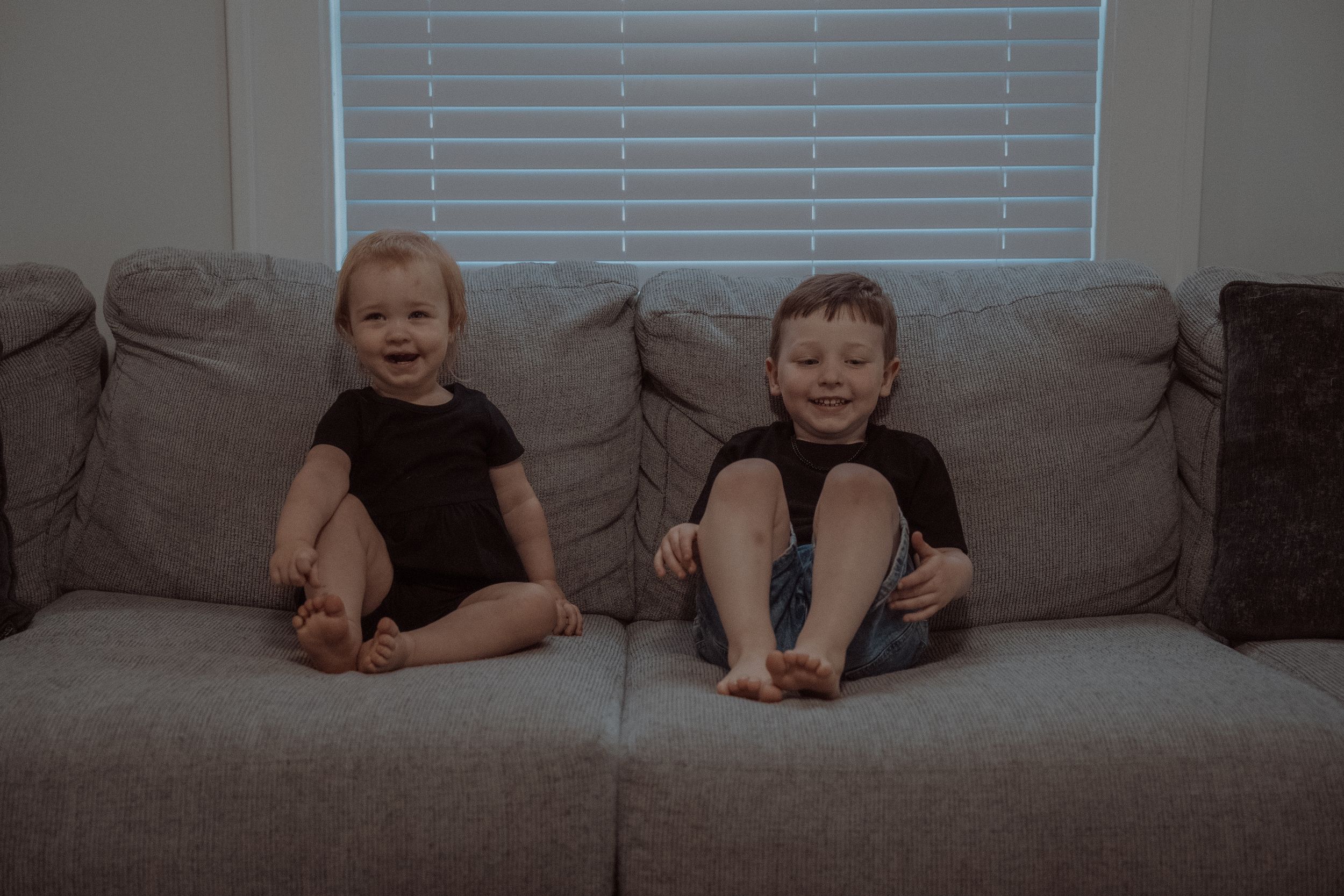 Two children in dark clothing sit together on a gray couch by a window with blinds.