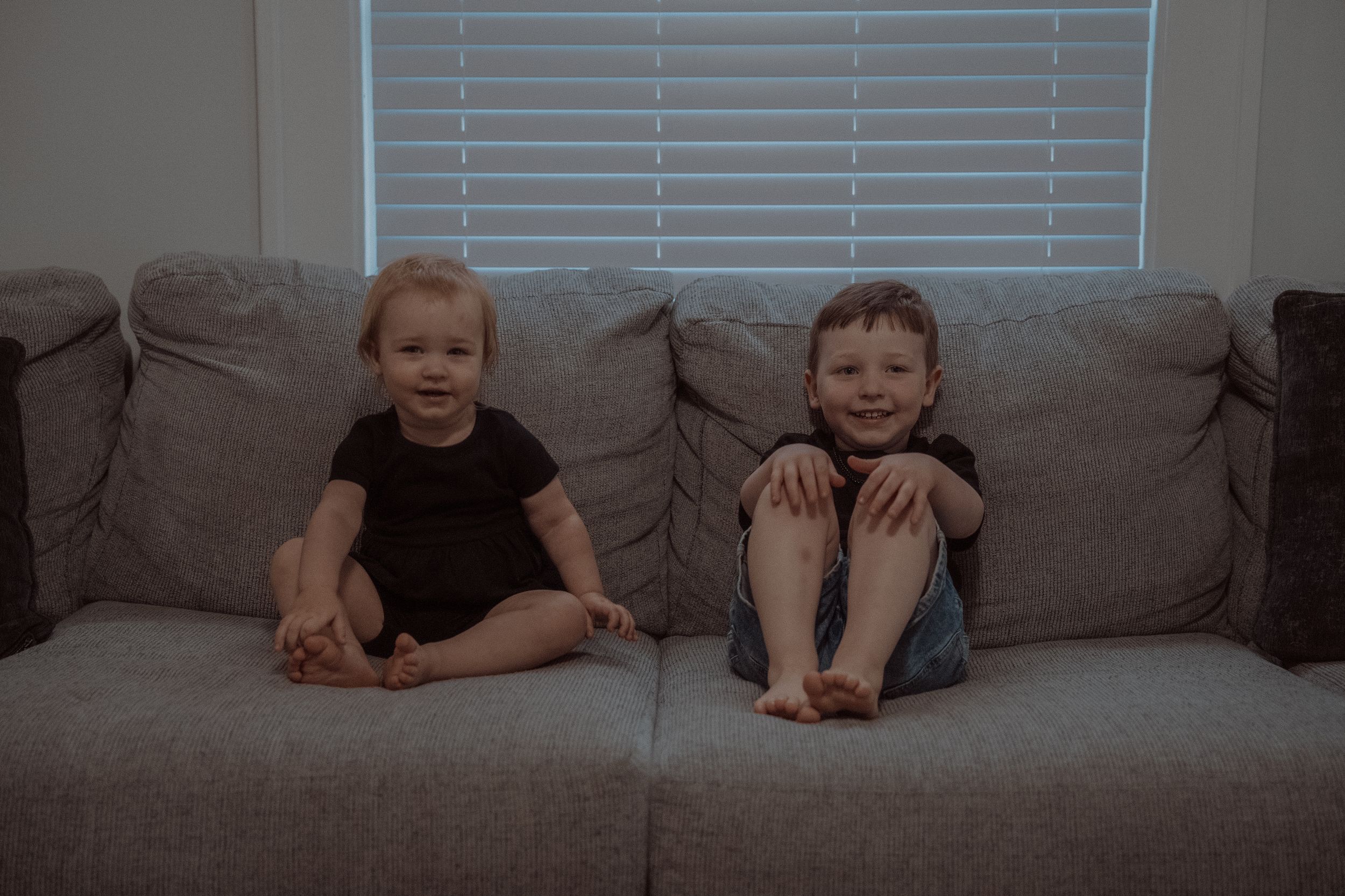 Two children sitting together on a gray couch in front of window blinds.