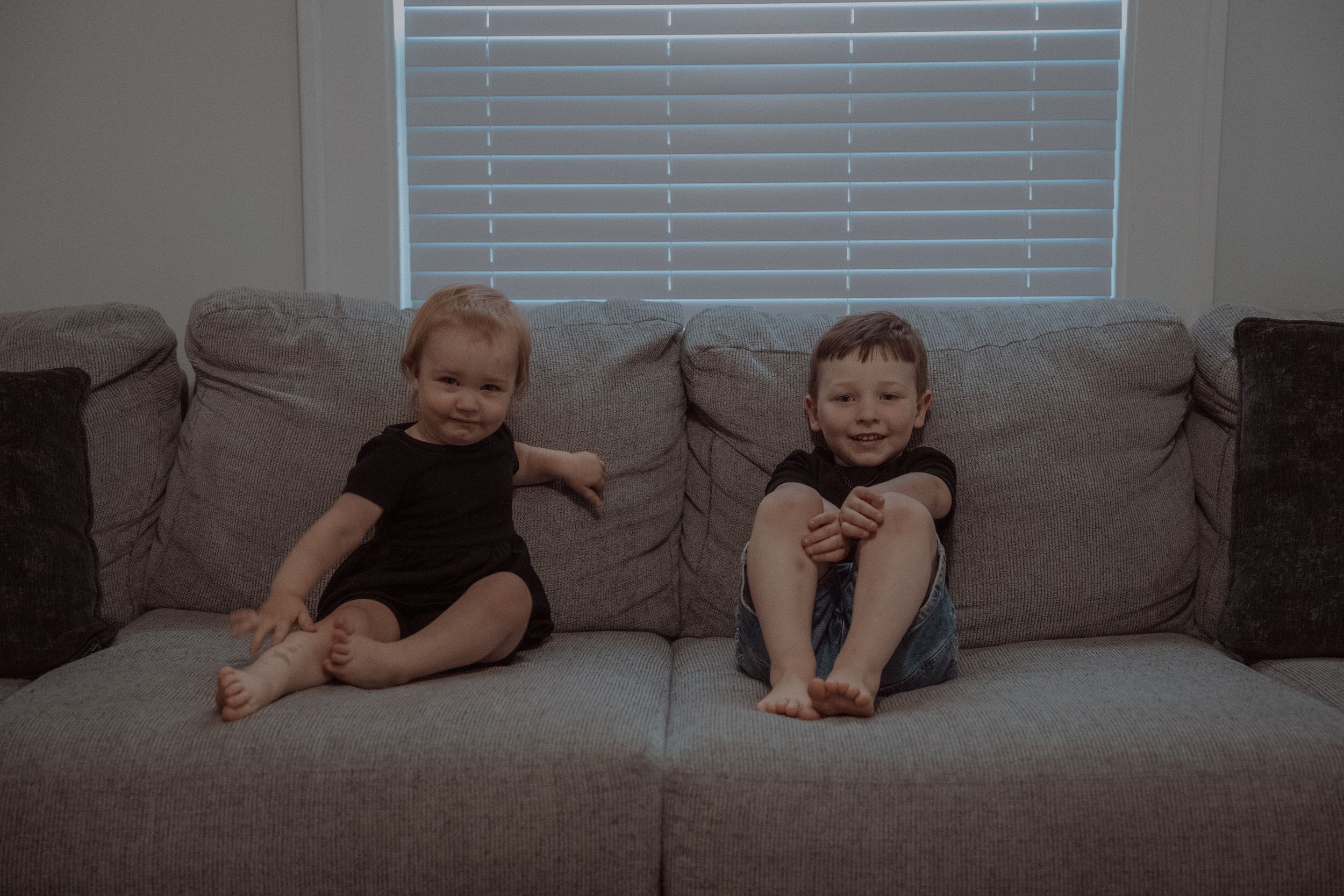 Two small children in dark outfits sit together on a gray couch beneath a window with blinds.