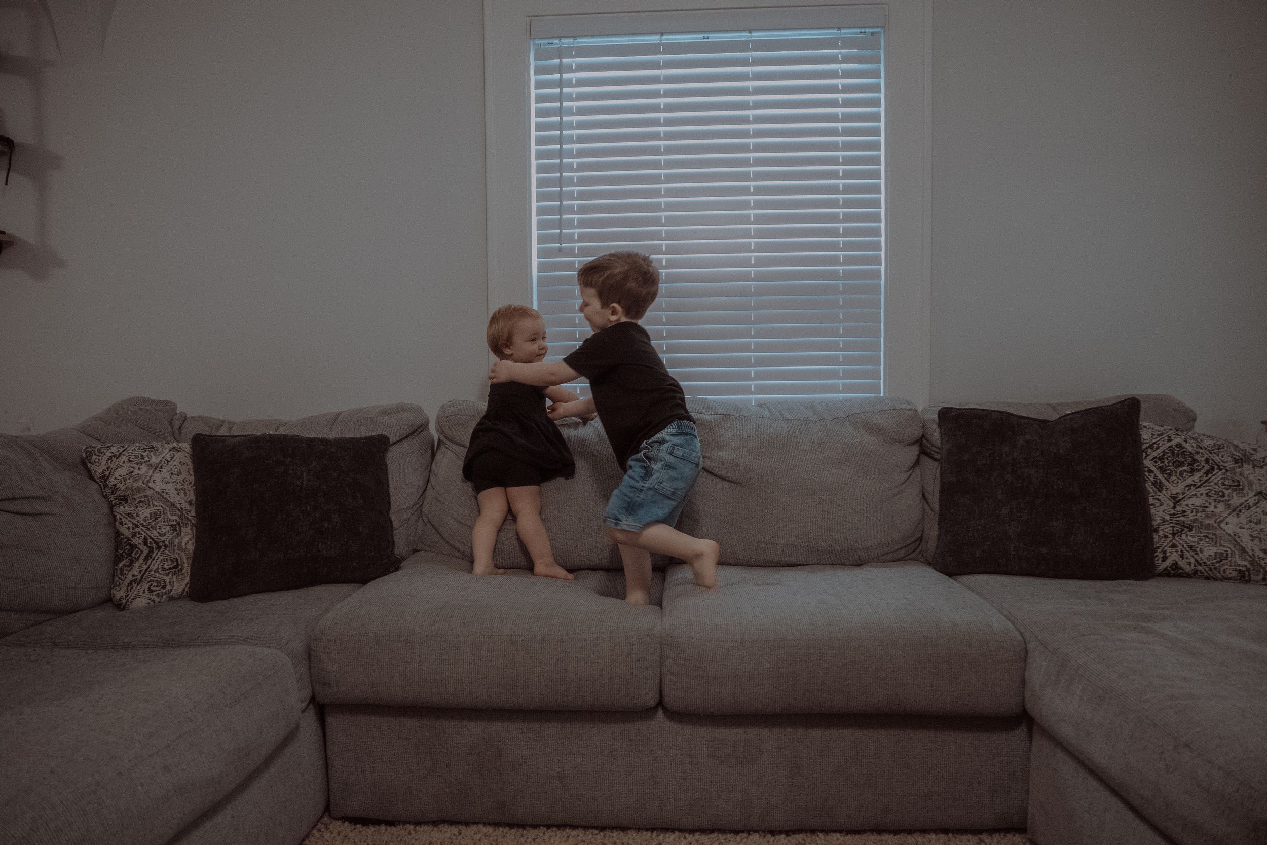 Two young children playing and jumping together on a gray sectional couch in a living room.
