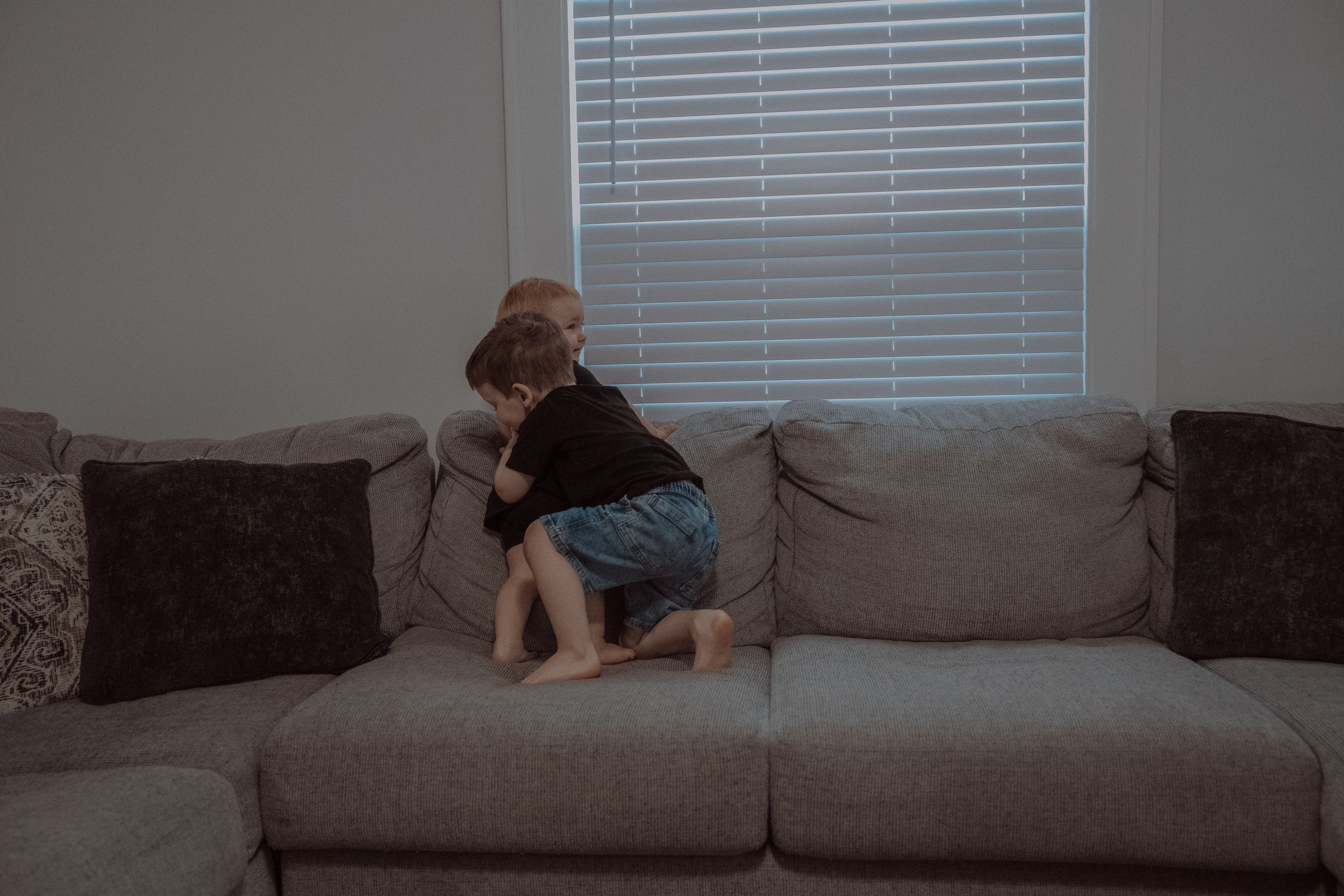 A young child sits on a gray sectional sofa with dark decorative pillows near a window with horizontal blinds.