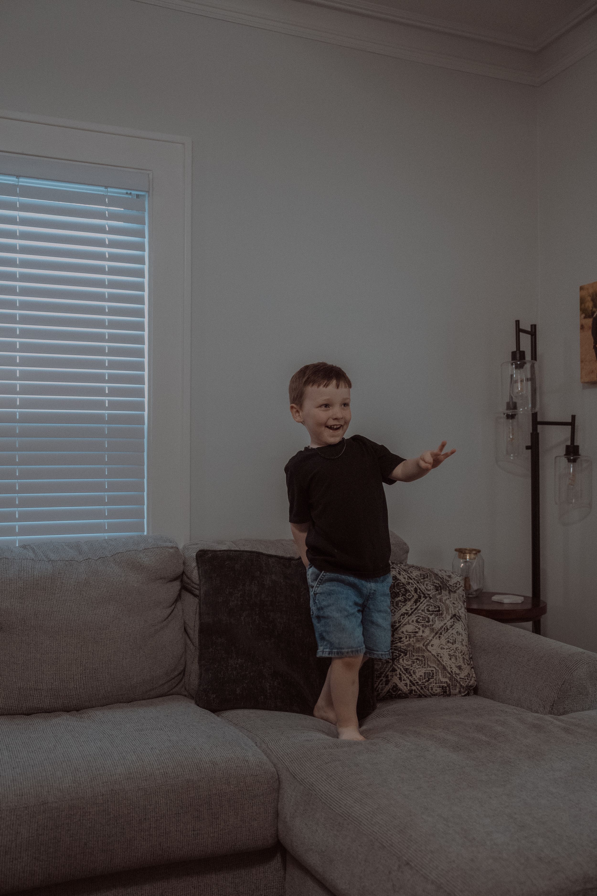 A small child plays and jumps on a gray couch in a modern living room with window blinds.