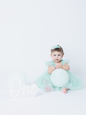 Baby in mint green tulle dress sits with balloon and 'one' text decoration on white background.