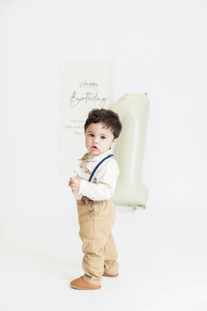 Small child wearing khaki pants with suspenders and a white shirt stands against light background.