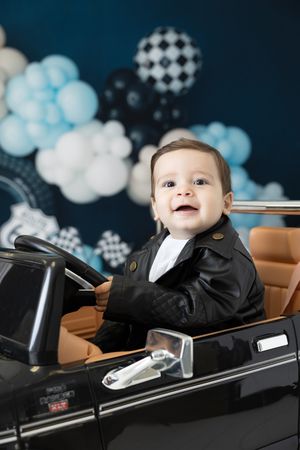 Smiling baby in leather jacket sitting in toy car with balloon backdrop.