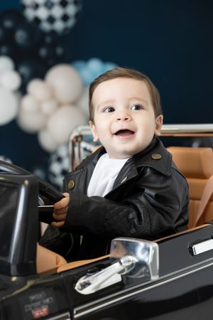 Smiling baby in black leather jacket sits in toy car with balloon backdrop.