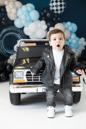 Toddler boy in leather jacket and jeans stands before toy SUV with blue and black balloon backdrop.