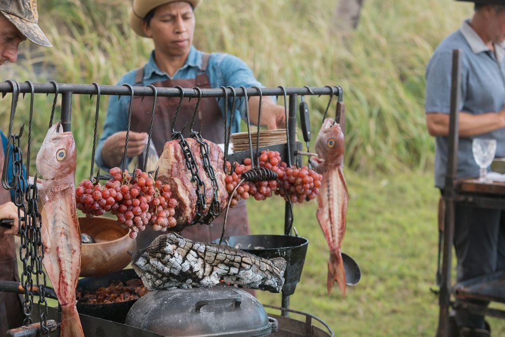 A rustic outdoor grill setup shows meat and sausages hanging over charcoal with fresh vegetables nearby.