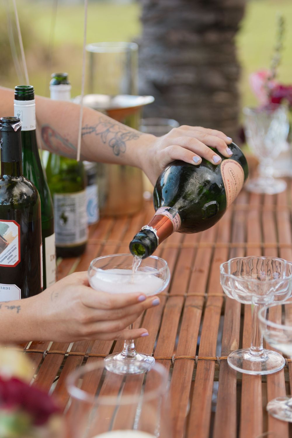 Someone pours champagne into a glass at an outdoor celebration table with multiple wine bottles and glassware.