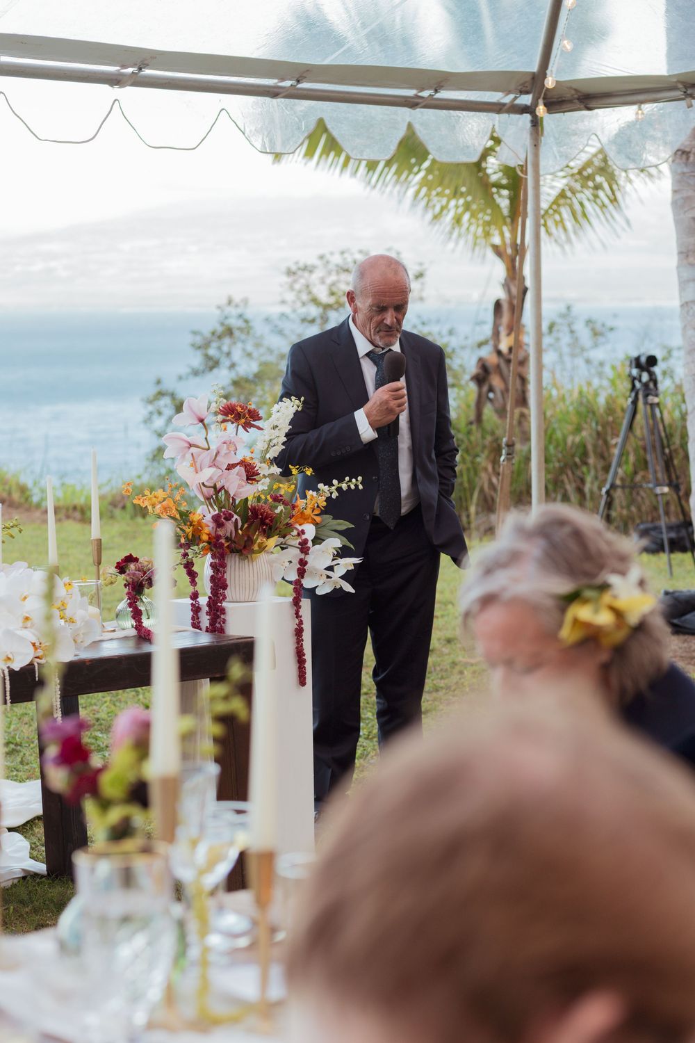 Outdoor wedding ceremony with speaker at podium overlooking ocean view under tent.