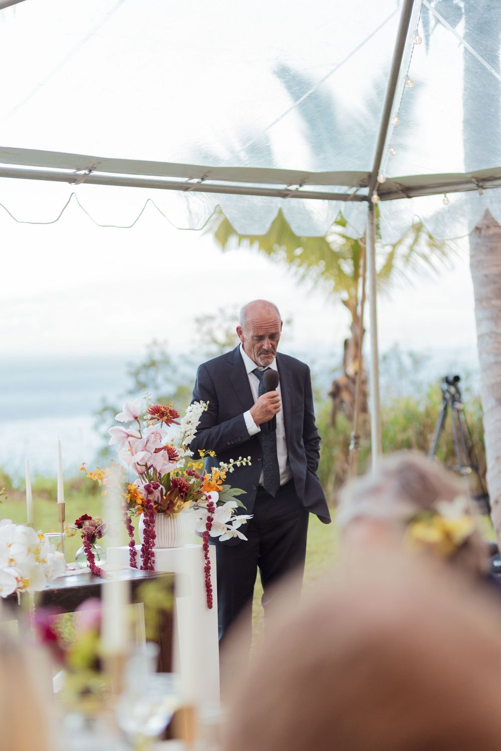 A speaker addresses guests under an outdoor wedding tent decorated with colorful floral arrangements.