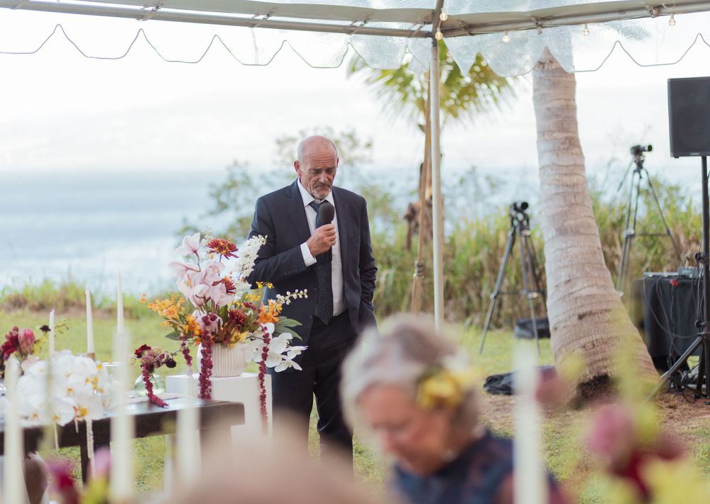 Speaker giving a speech at an outdoor beach wedding reception with palm trees and ocean views.