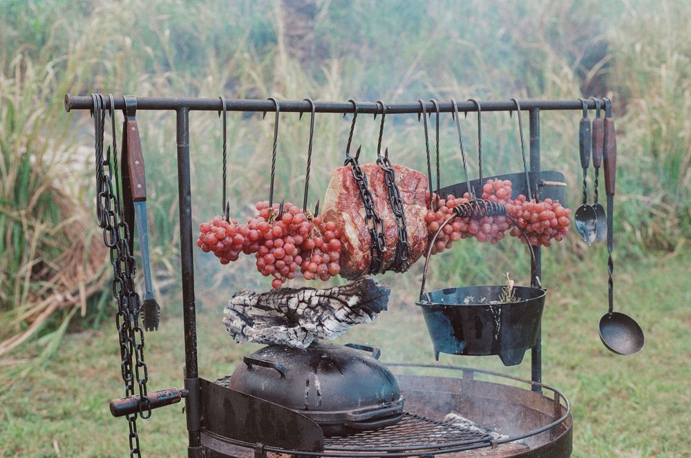 Old rustic cooking setup with dried meats and vegetables hanging over a metal grill in an outdoor setting.