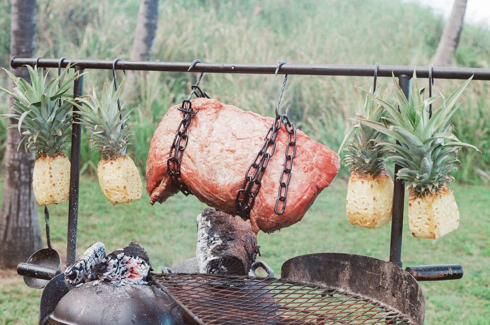A rustic outdoor barbecue setup with a large rotating metal spit roaster and decorative pineapple lanterns hanging above.