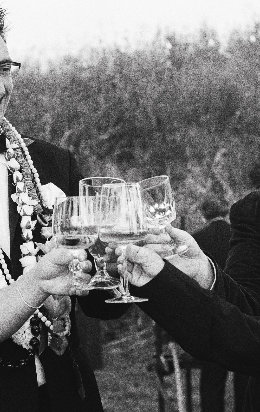 Two people in formal attire clink wine glasses together in black and white photography at an outdoor celebration.
