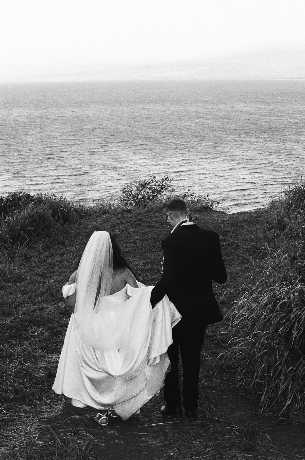 A bride in a white gown and groom in black suit stand at the edge of a cliff overlooking a misty ocean view.