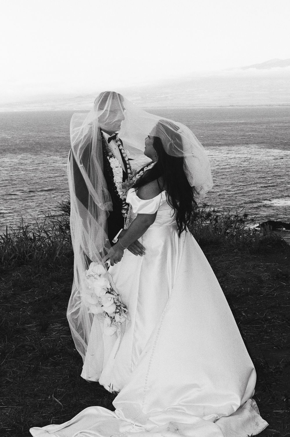 A romantic black and white wedding photo of a couple sharing a moment on a scenic coastal cliff overlooking the ocean.