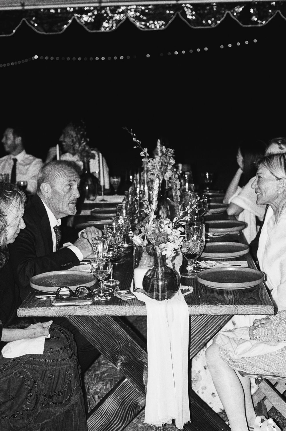 Black and white photo of a formal dinner party with table settings, flowers and candles under decorative string lights.