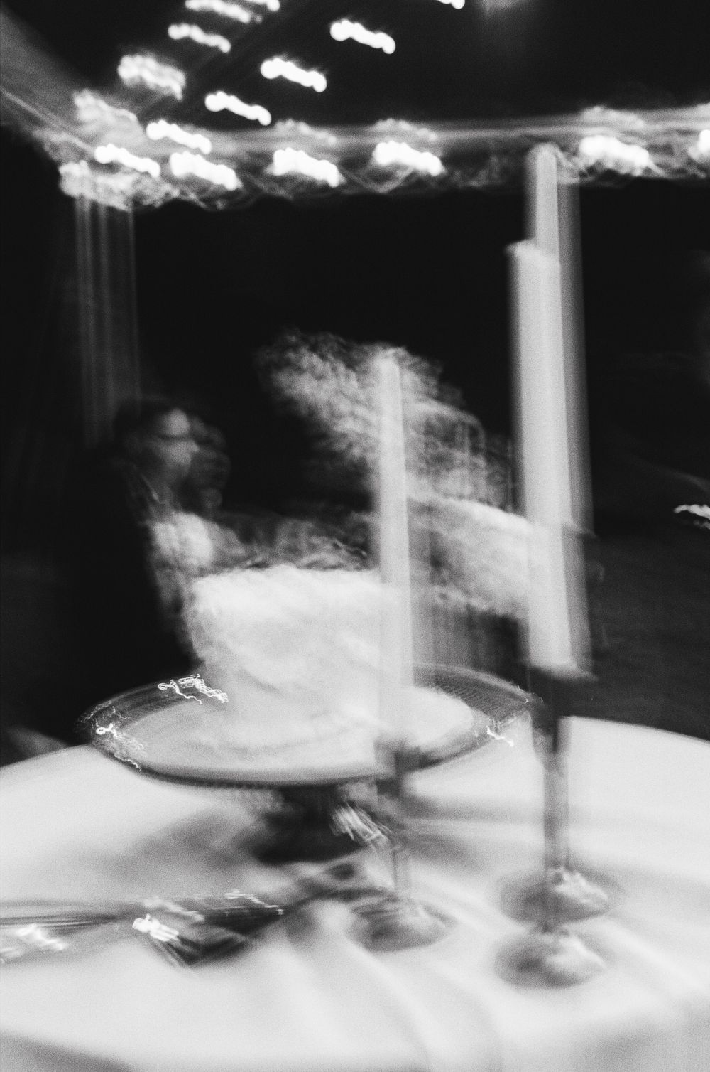 Motion-blurred artistic black and white photograph of a table setting with wine glasses and lit candles at night.