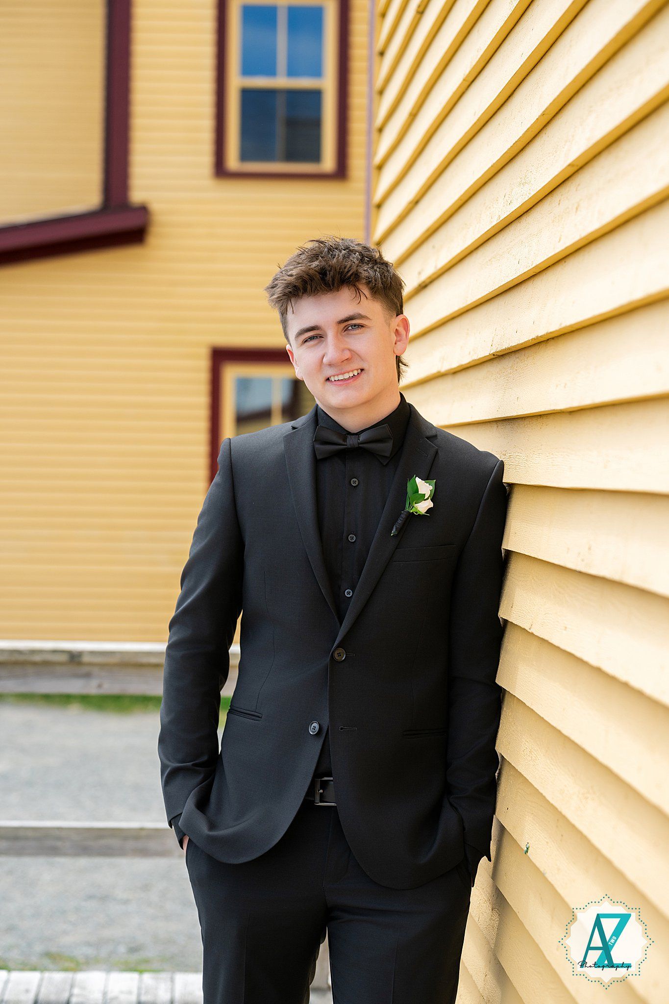 Young person in black formal tuxedo with boutonniere poses against yellow siding of house.