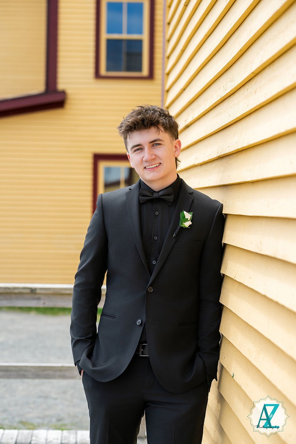 Young person in black formal tuxedo with boutonniere poses against yellow siding of house.