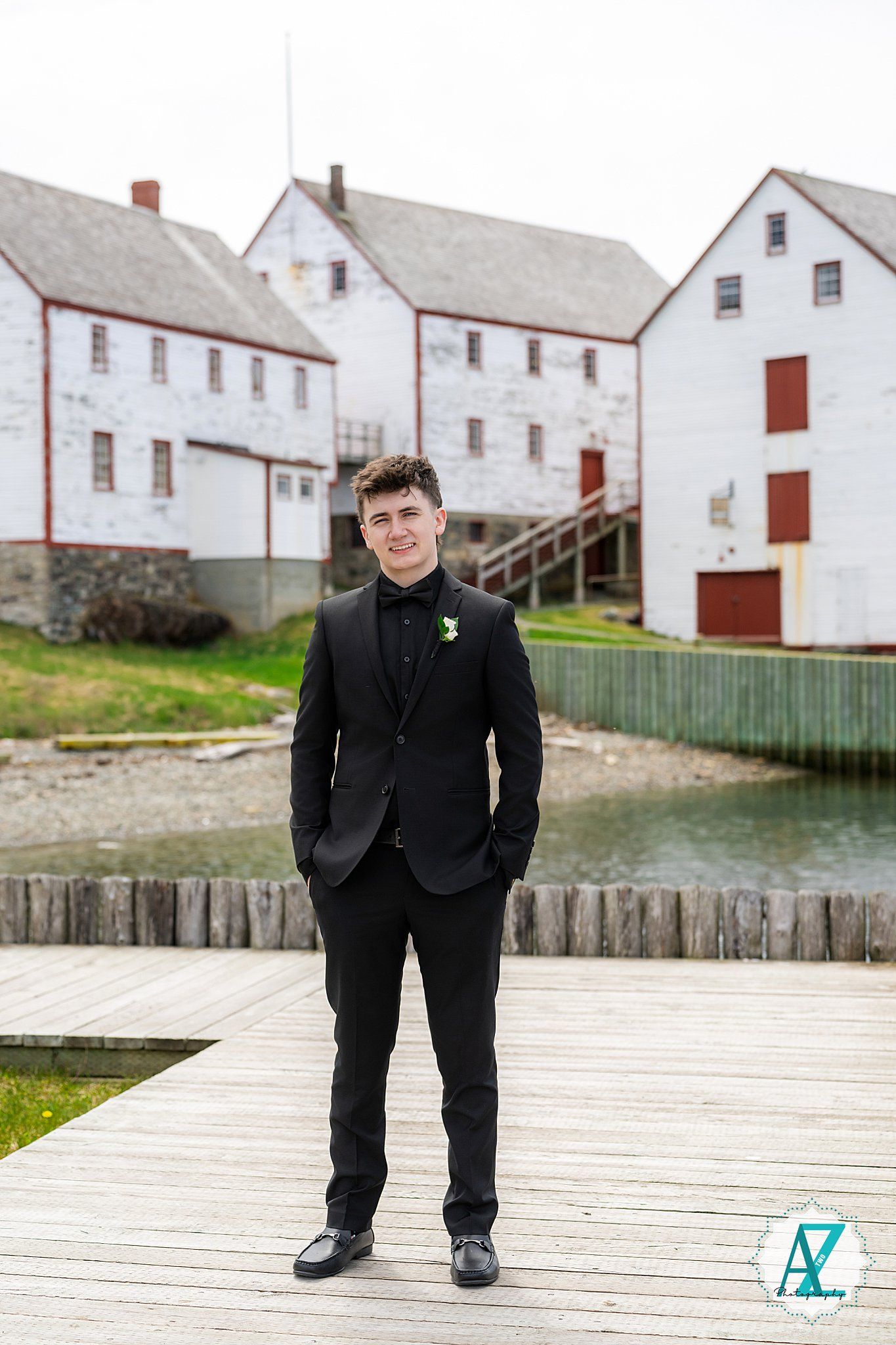 A person in a formal black suit stands on a wooden dock with historic white waterfront buildings in the background.