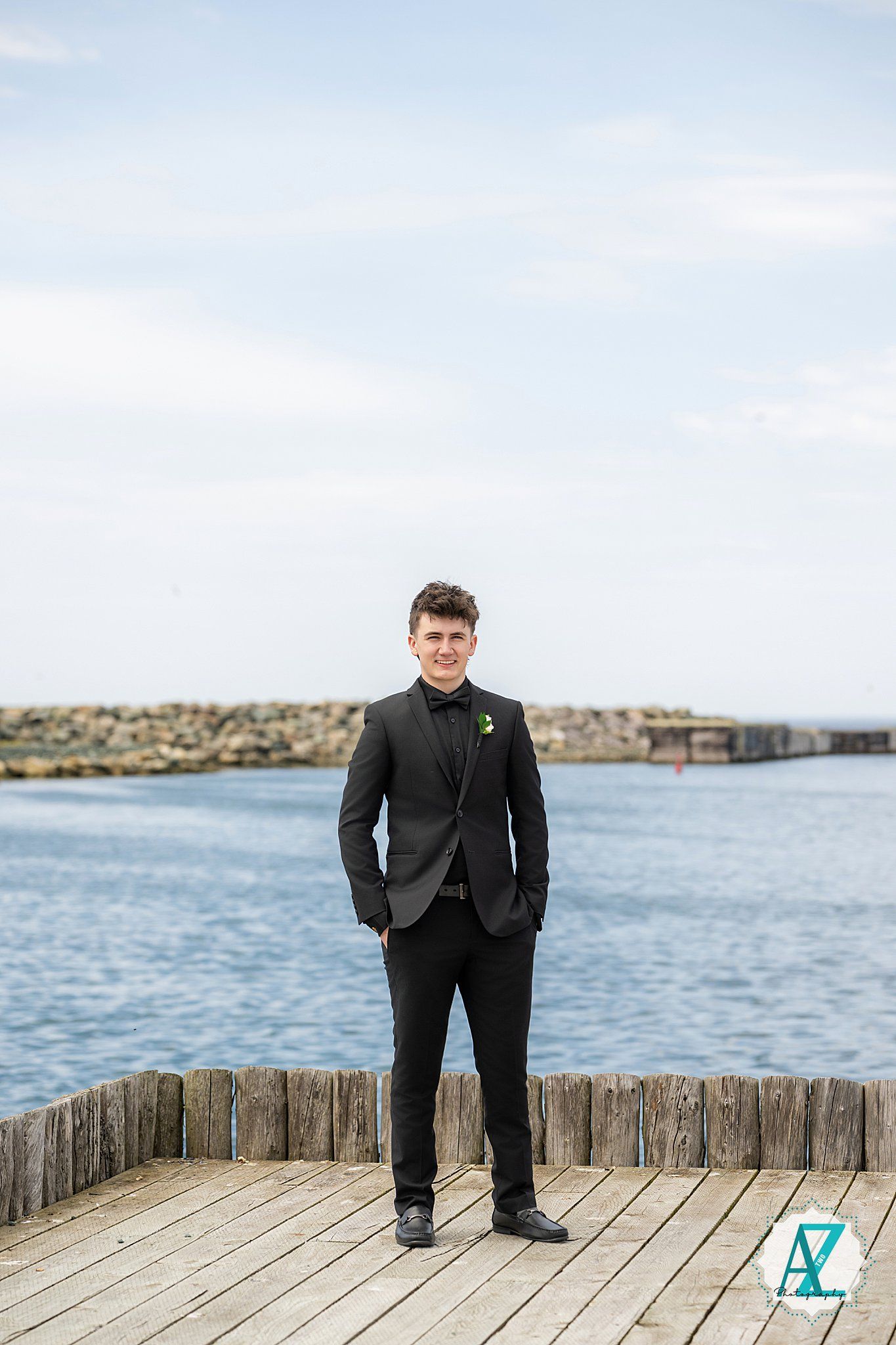 A person in a dark suit stands on a wooden pier by the water on a bright day.