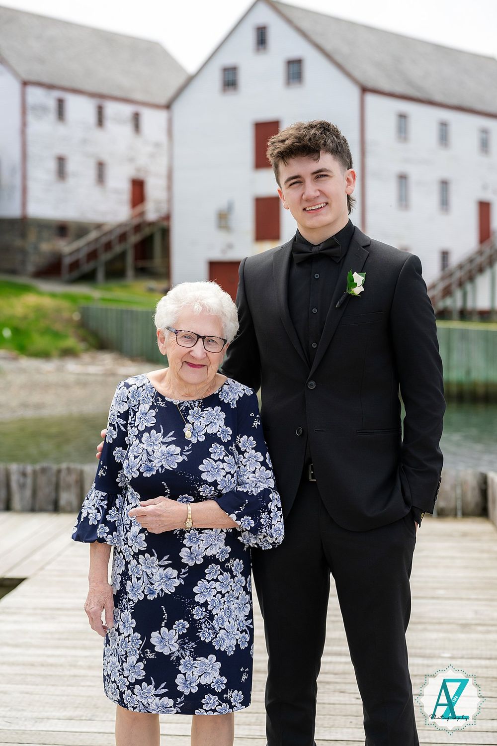 A formal portrait of two people wearing black and floral attire standing on a dock by white buildings.
