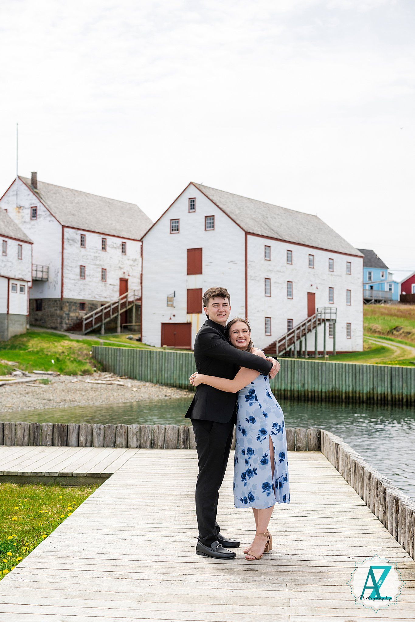 A couple embraces on a waterfront boardwalk near historic white buildings in a coastal town.