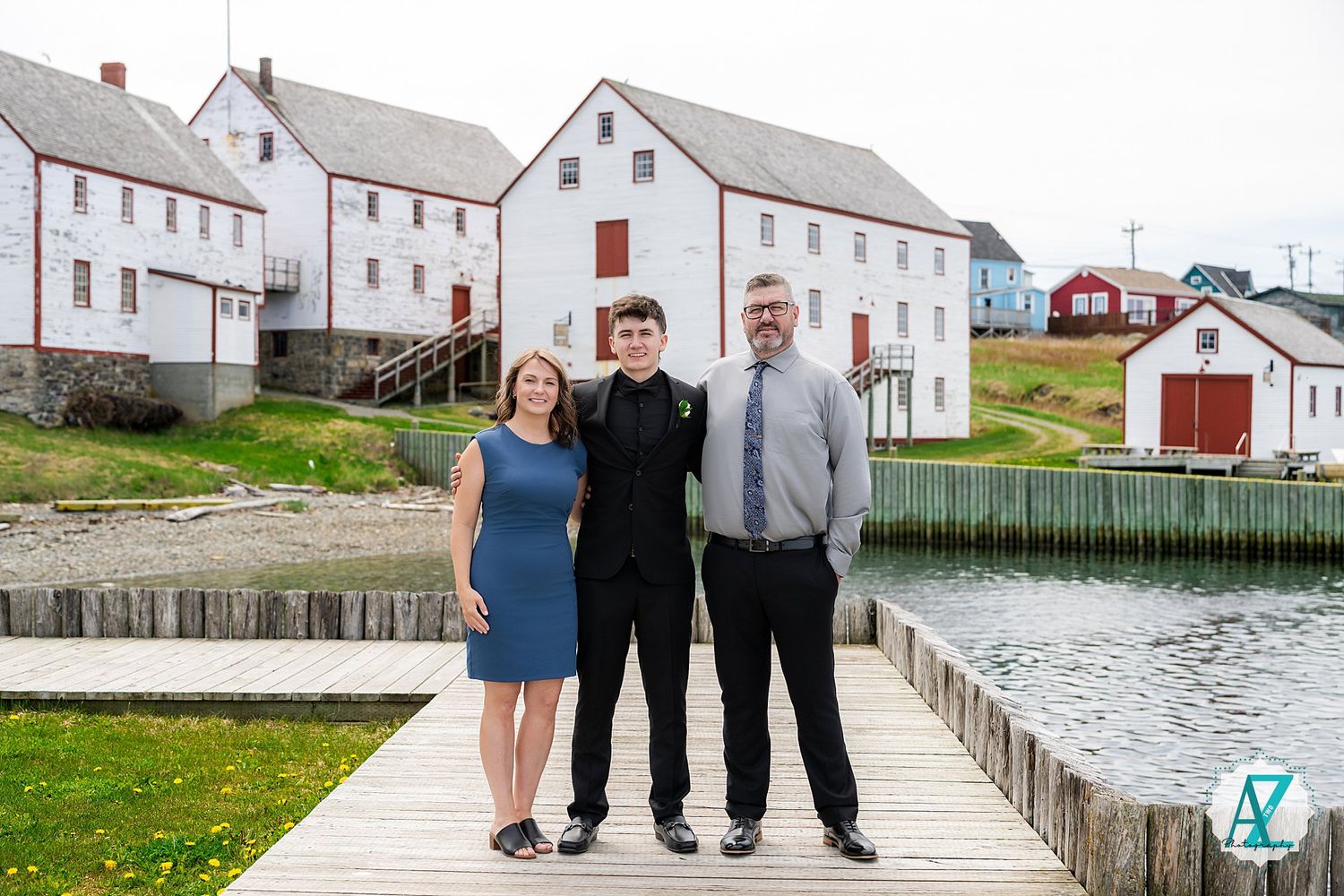 Three people stand on a wooden dock with white coastal buildings and water in the background in Maine.