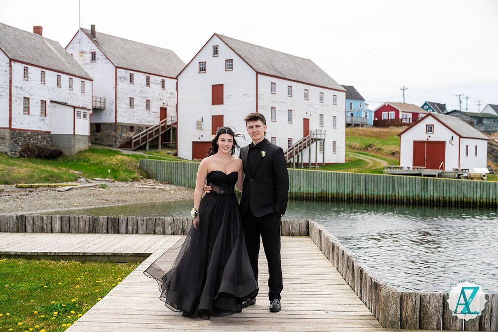 A couple poses together on a wooden boardwalk near white buildings and coastal waters in a scenic harbor setting.