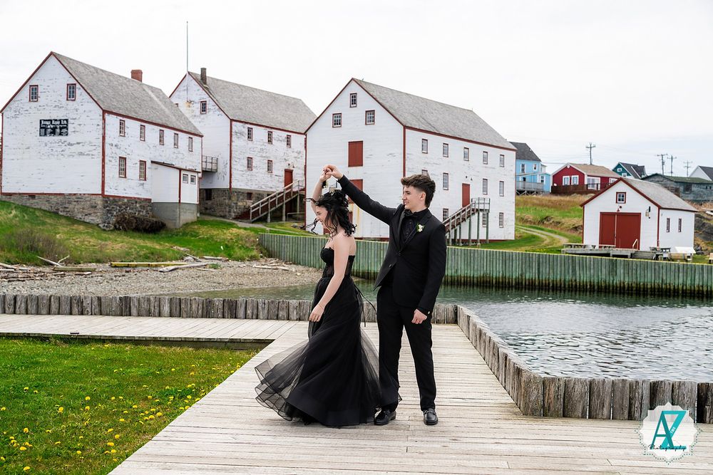 A couple stands on a wooden boardwalk near white buildings and water in a coastal setting.