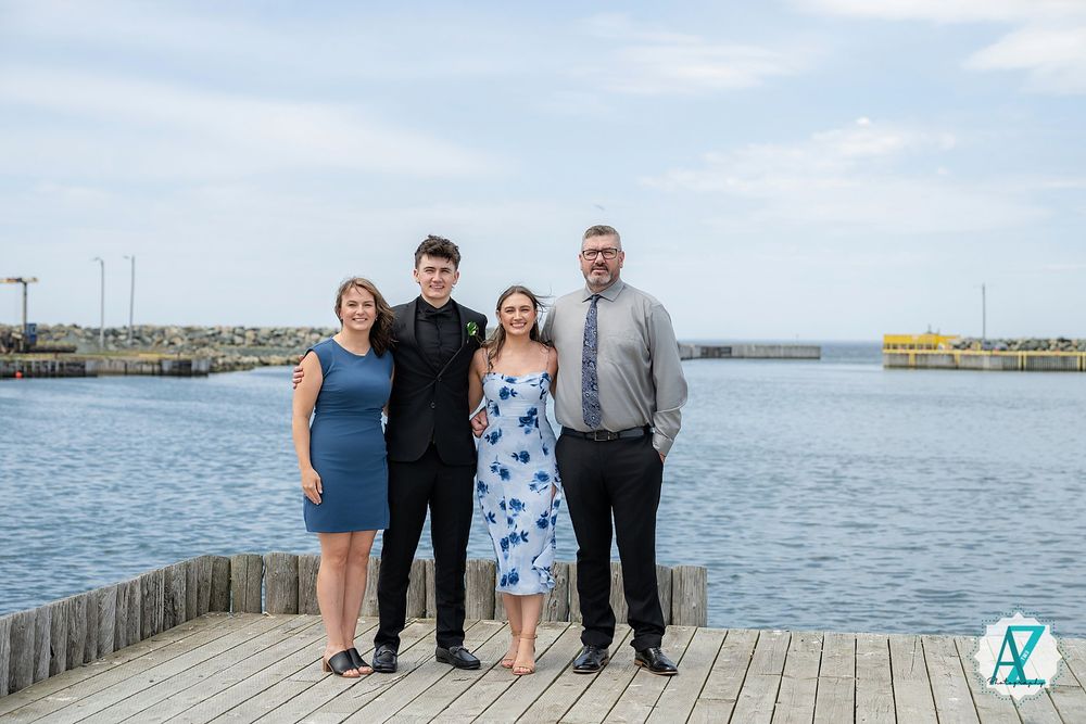 A group of four people in formal attire pose together on a wooden dock with water and boats in background.