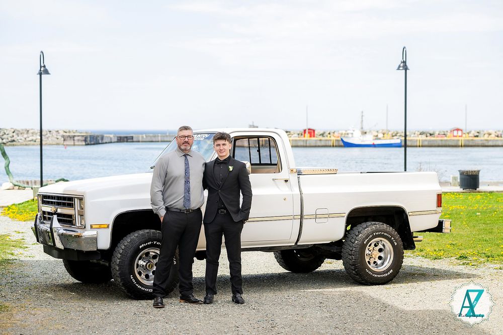 Two people pose beside a lifted silver vintage pickup truck near a harbor.