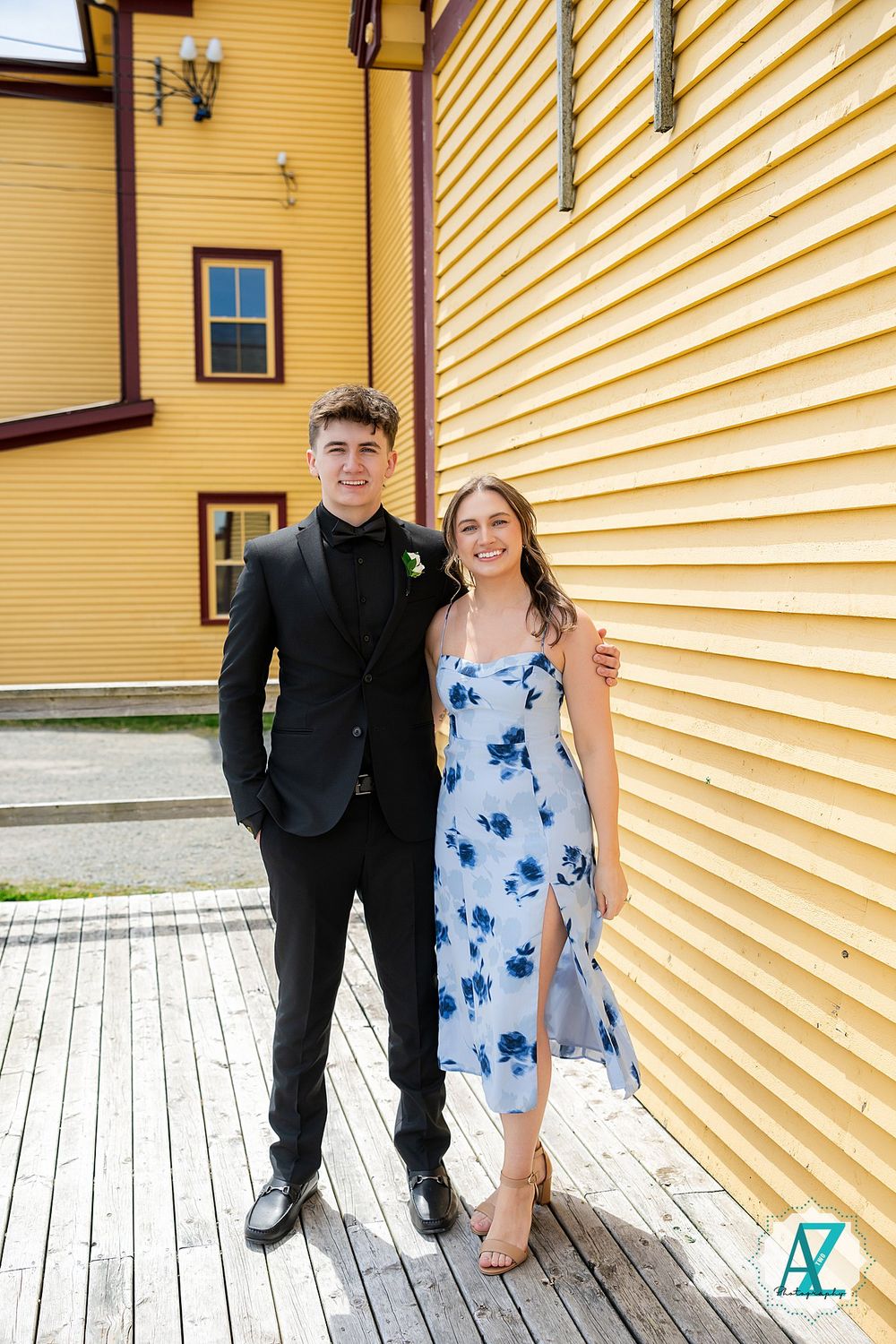 A couple stands in formal attire against a yellow building exterior with brown trim on a wooden deck.
