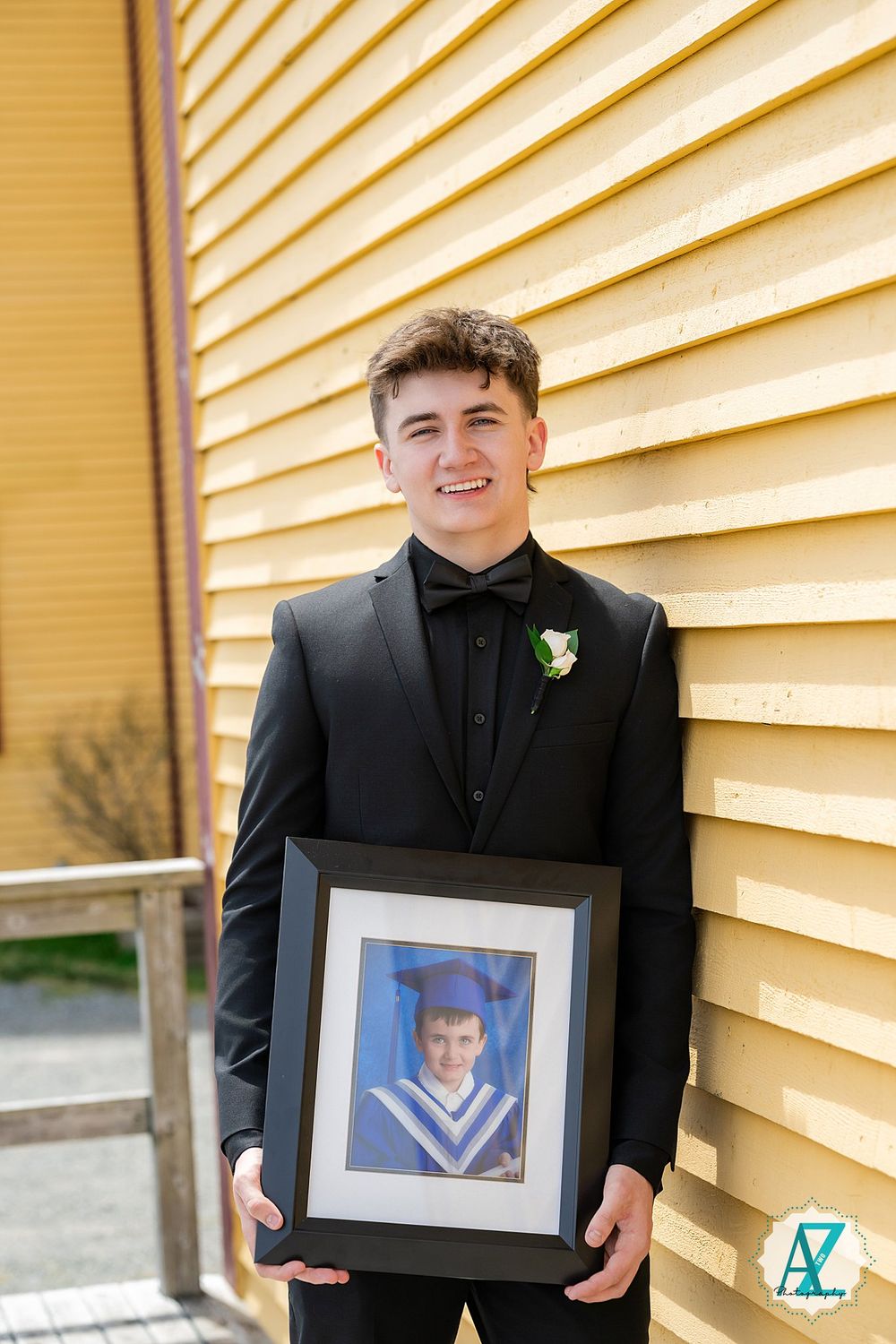 A graduate in a black tuxedo holds a framed childhood graduation photo against a yellow siding background.