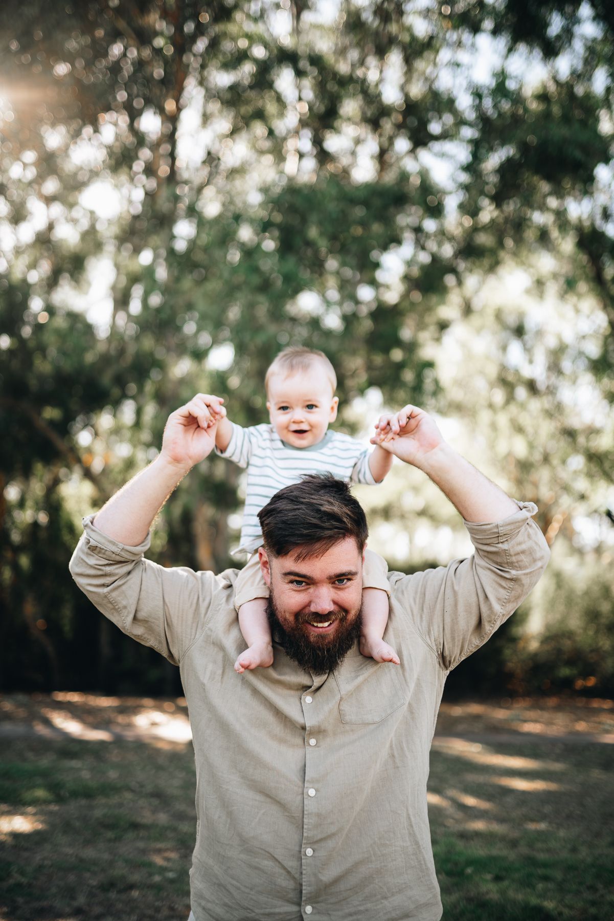 A joyful family moment captured outdoors with sunlight filtering through trees in the background taken by Melbourne Family photographer.
