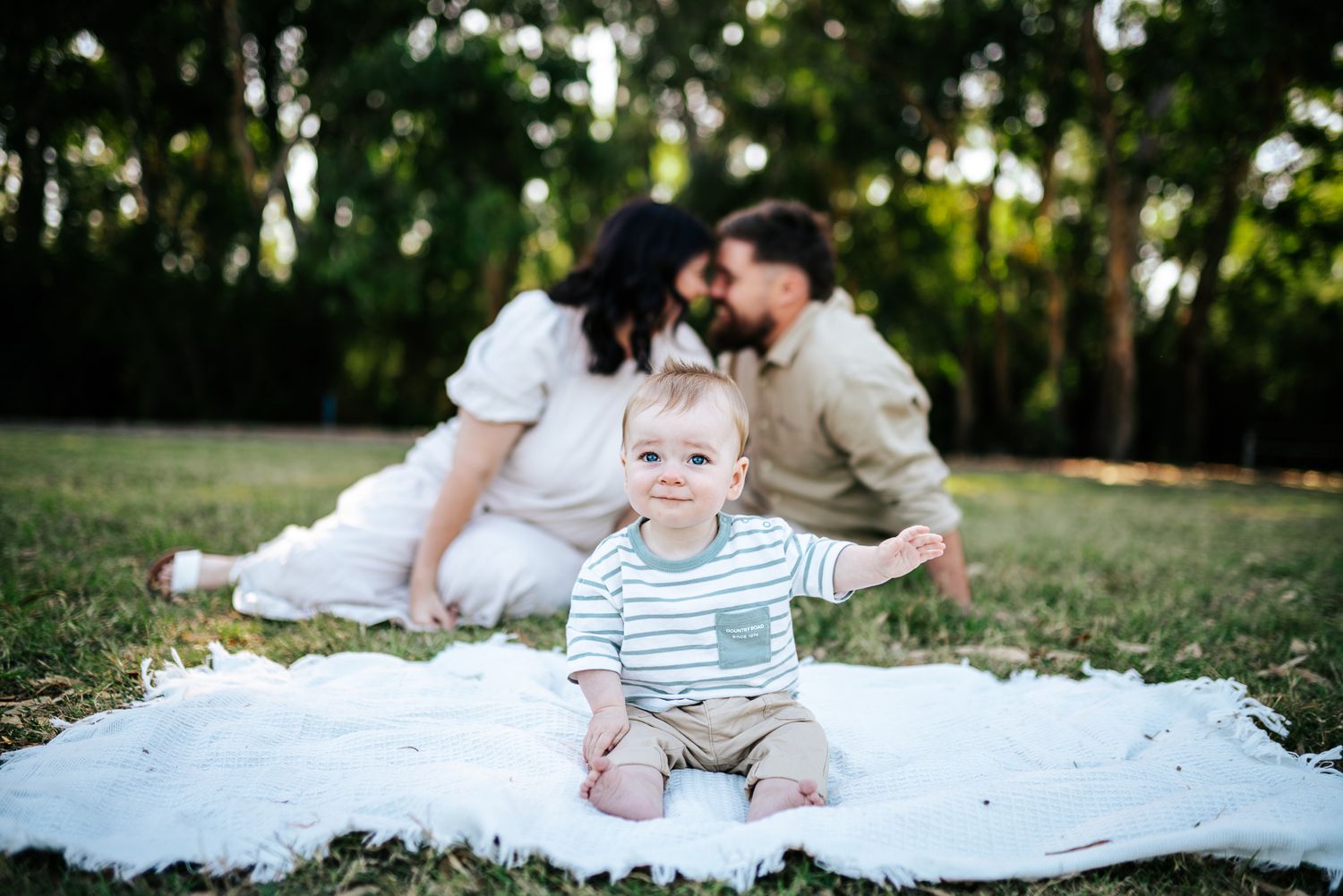 Baby sits on white blanket in grass while parents rest on picnic blanket in garden setting taken by Melbourne family photographer.