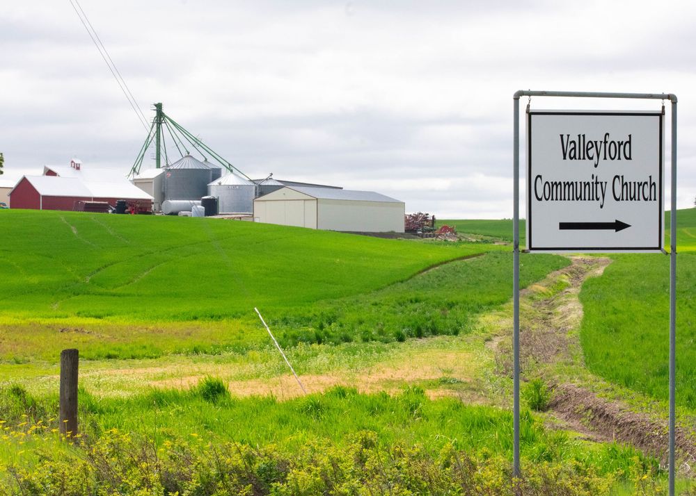 Sign directing to Valleyford Community Church beside green rolling farmland with grain silos in background.