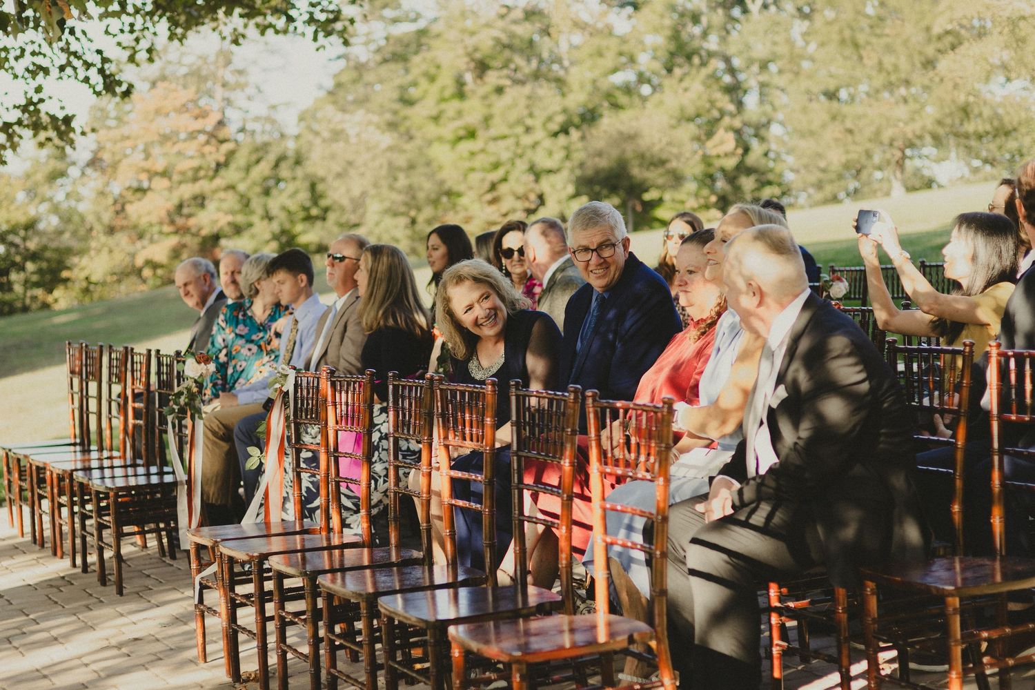Wedding ceremony guests sitting in wooden chairs during an outdoor autumn celebration with sunlit trees in the background.