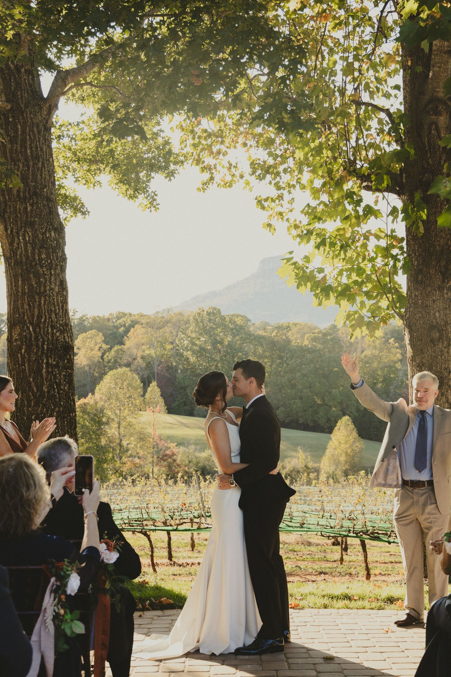 A couple embraces during their wedding ceremony at a scenic vineyard with mountains in the background at sunset.
