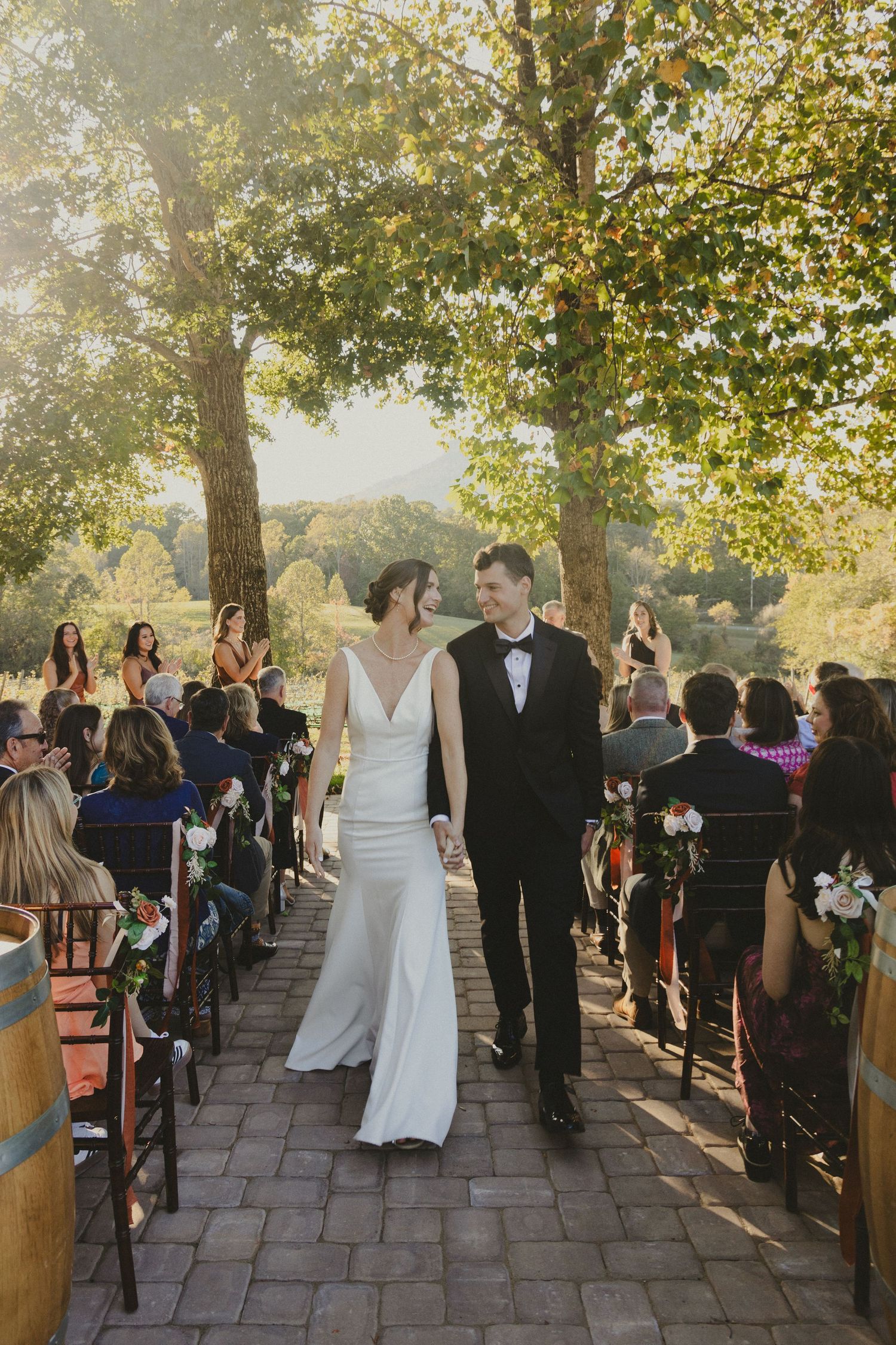 Newlyweds walk down a tree-lined stone path aisle while wedding guests look on during an autumn outdoor ceremony.